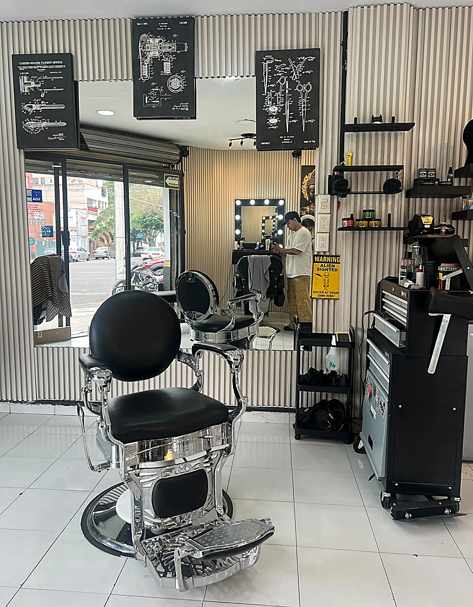 Interior de MG Barbershop en Torres Adalid: silla de barbero cromada, muro de listones y cuadros de patentes antiguas.